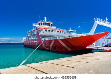 Ferry At Shore On Thassos Island, Greece - Waiting For The Tourists To Come Aboard. Red Boat Docked With Crystal Clear Waters And Blue Sky.