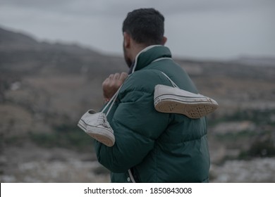 Fes/Morocco - November 06, 2020; Man Wearing Green Winter Jacket Looking In The Opposite Direction Of The Camera While Hanging A Pair Of White Converse Shoes On His Shoulders During A Cloudy Day