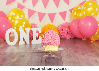 Festive background decoration for birthday celebration with pink girl cupcake or cake on glass stand, letters saying one and pink red yellow balloons in studio. Cake-smash first year concept.