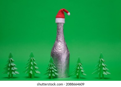 Festive Christmas Scene With Champagne Bottle And Green Origami Fir Tree. New Year And Christmas Card On A Green Background. New Year Background With Paper Fir Tree. Bottle Of Champagne With Santa Hat