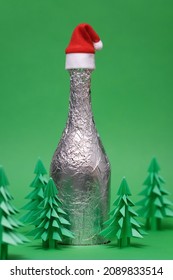 Festive Christmas Scene With Champagne Bottle And Green Origami Fir Tree. New Year And Christmas Card On A Green Background. New Year Background With Paper Fir Tree. Bottle Of Champagne With Santa Hat