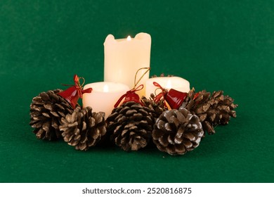 A festive Christmas table centerpiece featuring lit candles surrounded by pinecones and red bells on a green background - Powered by Shutterstock