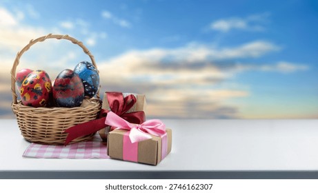 Festive composition featuring a wicker basket with hand-painted Easter eggs and wrapped gift boxes on a white tabletop against a blurred sky background