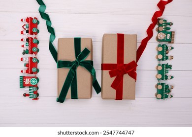 Festive composition with two gift boxes wrapped in kraft paper and tied with red and green velvet ribbons, accompanied by Christmas-themed toy trains on a white wooden background.