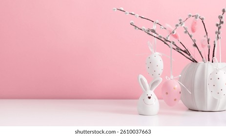 Festive Easter background. Vase with blooming pussy willow with Easter eggs and cute ceramic rabbit on white table against pink wall. Easter banner.