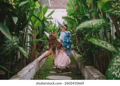 Festive Easter childrens game Egg hunt. Little brother and his older sister run along the path surrounded by tropical greenery in search of eggs, collect them in basket and have fun. Family traditions