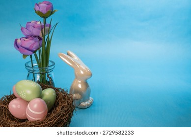 festive Easter composition with a ceramic bunny, a nest filled with pastel pink and green eggs, and a small glass vase of purple flowers against a solid blue background.