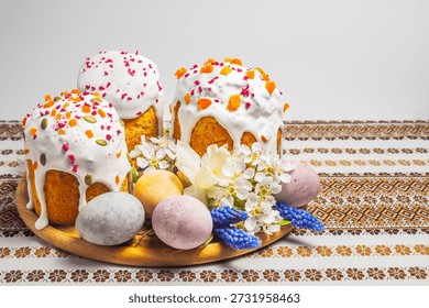 A festive Easter table setting features elaborately decorated kulich cakes surrounded by colorful eggs and delicate spring blossoms on a patterned tablecloth