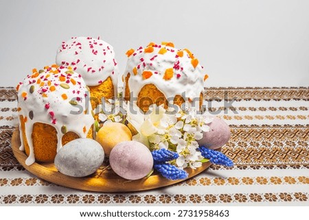 A festive Easter table setting features elaborately decorated kulich cakes surrounded by colorful eggs and delicate spring blossoms on a patterned tablecloth