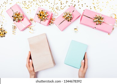 Festive Flat Lay Composition Female's Hands Holding Plain Gift Bag And Box Among Bright Presents And Ribbons On White Table, Top View.