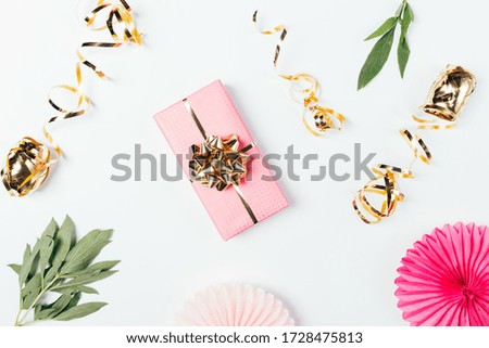 Festive flat lay composition of pink gift box with golden bow next to ribbons and decorations on white table.