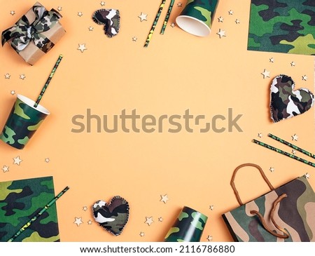 Festive frame for Defender of the Fatherland Day, February 23. Party set with camouflage glasses, straws, napkins, hearts and gifts on yellow background with copy space for text.