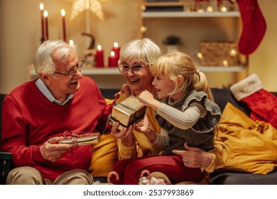 Festive grandparents and their little blonde granddaughter are sitting on a sofa, opening Christmas gifts together during Christmas - Powered by Shutterstock