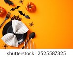 Festive Halloween table setting with black plate, striped napkin, cutlery, pumpkins, bats, and spiders on an orange background. Flat lay, top view.