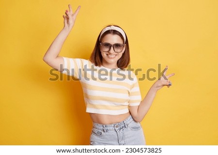 Festive happy woman wearing striped T-shirt hair band and sunglasses standing isolated over yellow background raised arms showing v sign celebrating victory.