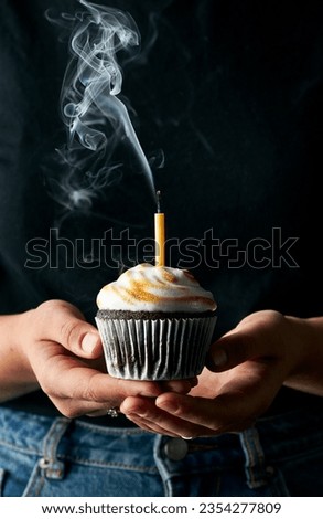 Festive image with a woman holding a chocolate cupcake topped with roasted meringue that has just blown out candle that is still smoking. Dark black background. Vertical image. 