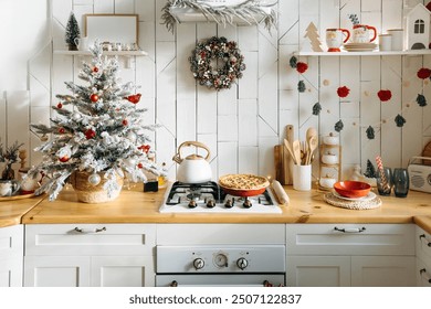 Festive kitchen featuring a small frosted Christmas tree, a pinecone wreath, red and white ornaments, and a freshly baked pie on the counter, creating a warm holiday atmosphere - Powered by Shutterstock