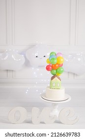 Festive location with the first birthday cake white background with balloons in the form of a cloud, blue lights. On the cake is a gingerbread house and colorful balloons, letters ONE.