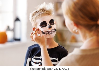 Festive Makeup For Halloween. Woman Doing Skeleton Make-up    For A Little Cheerful Boy In Costume While Preparing Holiday At Home.