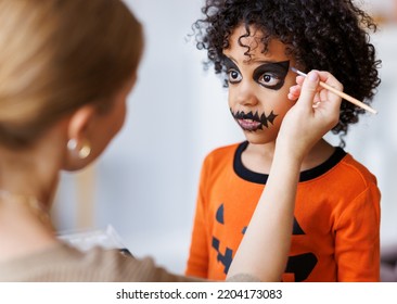 Festive Makeup For Halloween. Woman Doing Pumpkin Make-up  For Cheerful Ethnic Curly Boy In Costume While Preparing Holiday At Home.