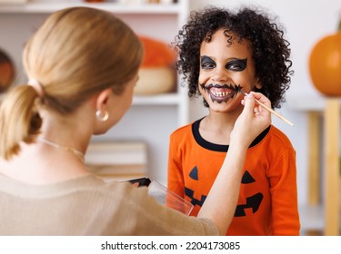 Festive Makeup For Halloween. Woman Doing Pumpkin Make-up  For Cheerful Ethnic Curly Boy In Costume While Preparing Holiday At Home.