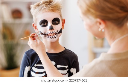Festive makeup for Halloween. Woman doing skeleton make-up    for a little cheerful boy in costume while preparing holiday at home. - Shutterstock ID 2209374601