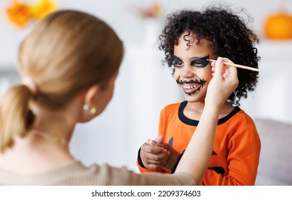 Festive Makeup For Halloween. Woman Doing Pumpkin Make-up  For Cheerful Ethnic Curly Boy In Costume While Preparing Holiday At Home.