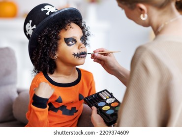 Festive Makeup For Halloween. Woman Doing Pumpkin Make-up  For Cheerful Ethnic Curly Boy In Costume While Preparing Holiday At Home.