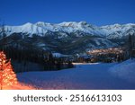 A festive mountain village illuminated by warm holiday lights, nestled below snow-covered peaks. The scene captures the peaceful beauty of a winter wonderland, perfect for holiday and ski adventures.