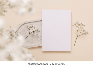 Festive paper greeting card on ceramic tray and gentle gypsophila flowers on beige background, top view. Mockup paper card for design, print, wedding and invitation