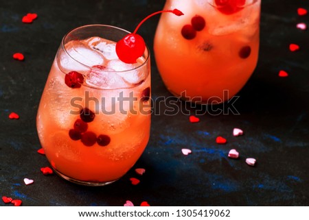 Festive pink cold alcoholic cocktail with red maraschino cherries for Valentines day, two glasses, black background with sweet red hearts, selective focus