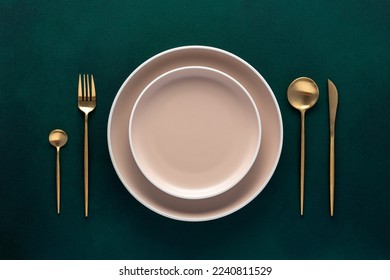 Festive place setting with beige dishes. Empty plate and gold cutlery on dark green background. Dining table in luxury restaurant. Card or menu template, flat design. Tableware, crockery. Top view.