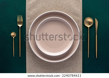 Festive place setting with beige napkin. Empty plates and gold cutlery on dark green background. Top view. Dining table in luxury restaurant. Card or menu template, flat design. Tableware, crockery.