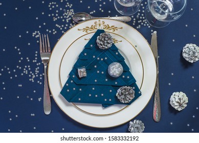 Festive Place Setting For Christmas Or New Year Holiday Dinner. Blue Napkin Shape Christmas Tree On White Plates, Beautiful Cutlery And Chrismtas Decorations On The Dark Blue Background. Flat Lay