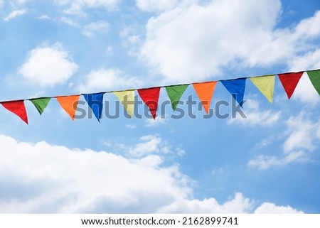 Festive ribbon of multicolored triangle flags on the white clouds and blue sky background. Celebration party, holiday fair, festival decorations.