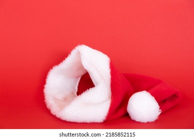 Festive Santa Claus Hat Lies Isolated On Red Background. Carnival Costume Accessory. Beautiful White Fur And Fluffy Pom Pom. Symbol Of Christmas And New Year. Holiday Mockup Design. Volume Side View.