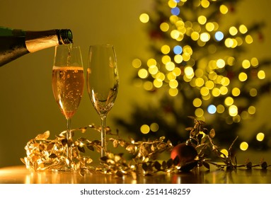 Festive scene of bottle pouring champagne into two glasses on table with gold decorations and red ornament ball.Christmas tree with blurred yellow bokeh lights background. Holidays celebration concept - Powered by Shutterstock