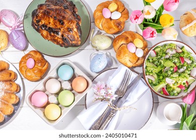 Festive served Easter dinner or brunch table, with Easter egg, tulip flower bouquet, traditional Easter foods - cake, cross buns, wreath bread, glazed ham and spring salad, white table background