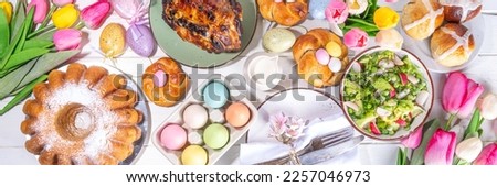 Festive served Easter dinner or brunch table, with Easter egg, tulip flower bouquet, traditional Easter foods - cake, cross buns, wreath bread, glazed ham and spring salad, white table background