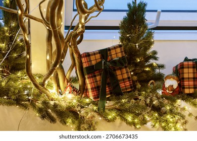 A festive setup featuring plaid-wrapped gift boxes with velvet ribbons, lush green branches, string lights, and miniature pine trees, creating a warm holiday ambiance.