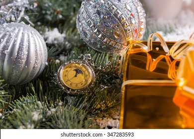 Festive Still Life Of Old Fashioned Pocket Watch On Snow Covered Evergreen Sprigs Decorated With Sparkling Silver Christmas Balls With Corner Of Gift Wrapped In Gold Paper With Bow