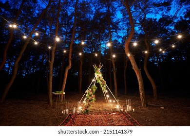 Festive string lights illumination on boho tipi arch decor on outdoor wedding ceremony venue in pine forest at night. Vintage string lights bulb garlands shining above chairs at summer rural wedding.