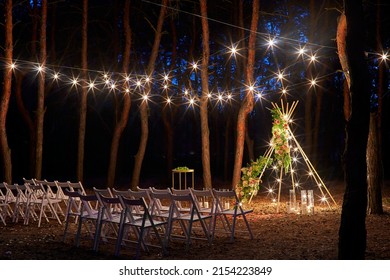 Festive string lights illumination on boho tipi arch decor on outdoor wedding ceremony venue in pine forest at night. Vintage string lights bulb garlands shining above chairs at summer rural wedding. - Powered by Shutterstock