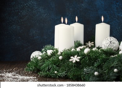 Festive stylish decor for holiday dinner at home. Christmas winter table setting with advent Xmas wreath, white candles, green branches and snowflakes on dark blue wooden background, copy space.