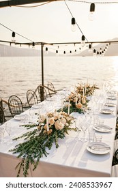 Festive table with bouquets of flowers and candles stands on a pier with garlands of light bulbs by the sea