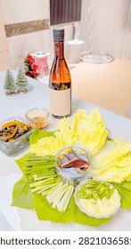 A festive table setting featuring a bottle of wine, fresh cabbage leaves, sliced scallions, and side dishes for a traditional Korean meal.