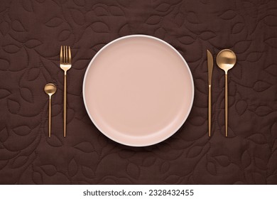 Festive table setting on dark tablecloth. Empty plate and gold cutlery on a brown background. Top view. Dining table in luxury restaurant. Card or menu template with space, flat design.