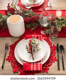 Festive Table Setting in Red, Snowflake Placemats, Crimson Table Runner, White Porcelain Plates with Pinecone Embellishment, Cutlery, Crystal Stemware, and a Centerpiece of Holly Berries and Candles.