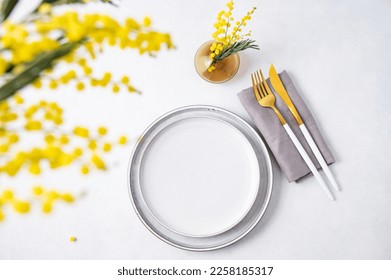 Festive table setting for the spring celebration of women's day, birthday or mother's day with yellow mimosa flowers on a light background with vase. Top view and copy space.