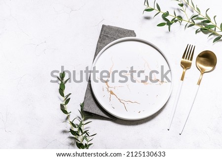 Festive table setting with white marble plate, golden cutlery set and fresh eucalyptus leaves on white rustic table top view. Copy space for your design.
